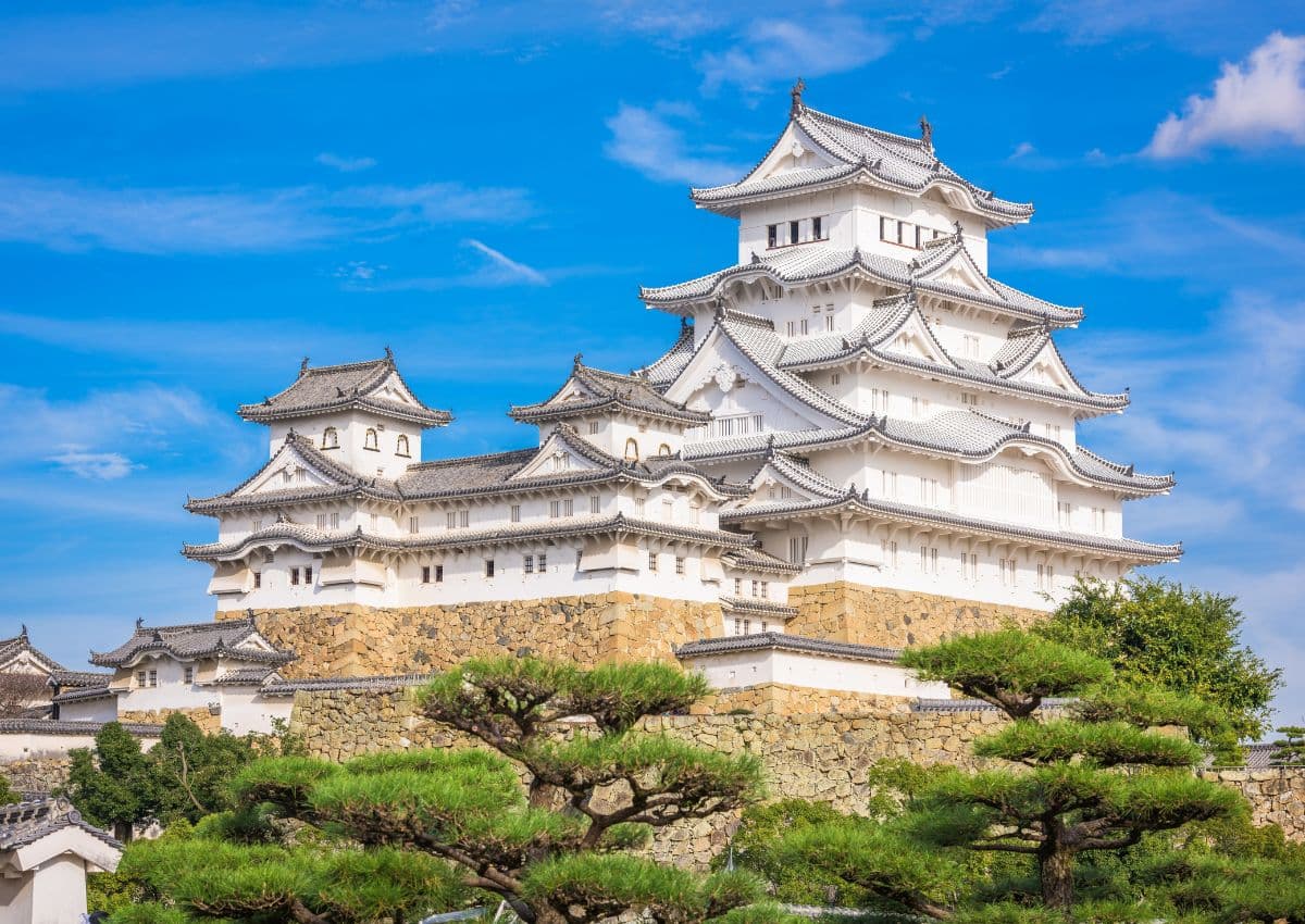 Burg Himeji, weiße japanische Burg und UNESCO-Welterbe, vor blauem Himmel und grünen Bäumen