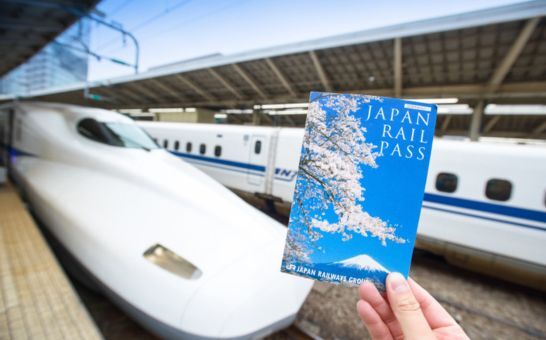Eine Hand hält ein Japan Rail Pass Ticket vor einem Shinkansen im Bahnhof von Tokio