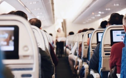 Innenraum eines großen Passagierflugzeugs mit sitzenden Menschen und einer Stewardess in Uniform, die den Gang entlanggeht