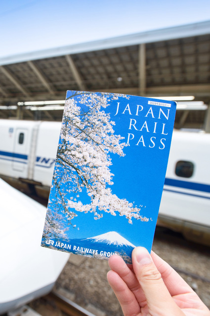 Eine Hand hält ein Japan Rail Pass Ticket vor einem Shinkansen im Bahnhof von Tokio